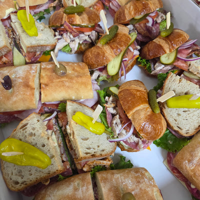 A close-up view of multiple croissant sub sandwiches cut into halves, arranged in a white container. The sandwiches are topped with melted cheese, sliced pickles, and mustard, and are held together with wooden skewers.
