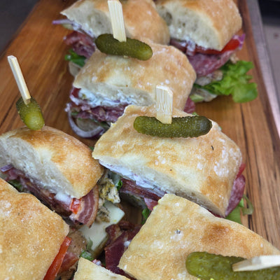 The Bigfoot Sandwich tray - a two-foot loaded ciabatta sandwich with deli meats, artisan cheese, pickles and fresh vegetables, cut into ten garnished pieces on a rustic board — Radical Gardens, Timmins