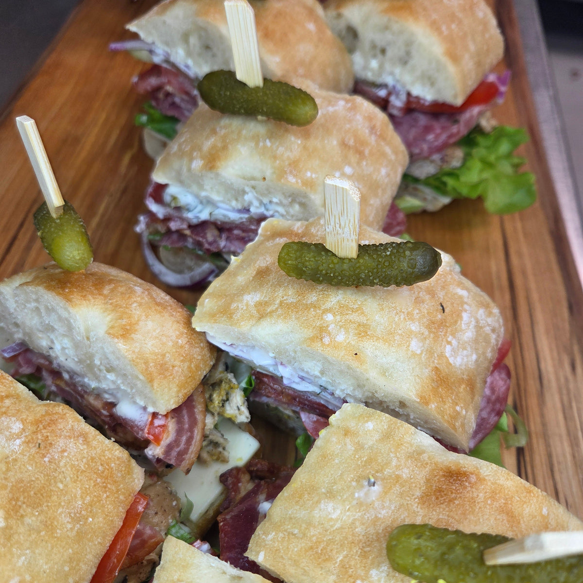 The Bigfoot Sandwich tray - a two-foot loaded ciabatta sandwich with deli meats, artisan cheese, pickles and fresh vegetables, cut into ten garnished pieces on a rustic board — Radical Gardens, Timmins